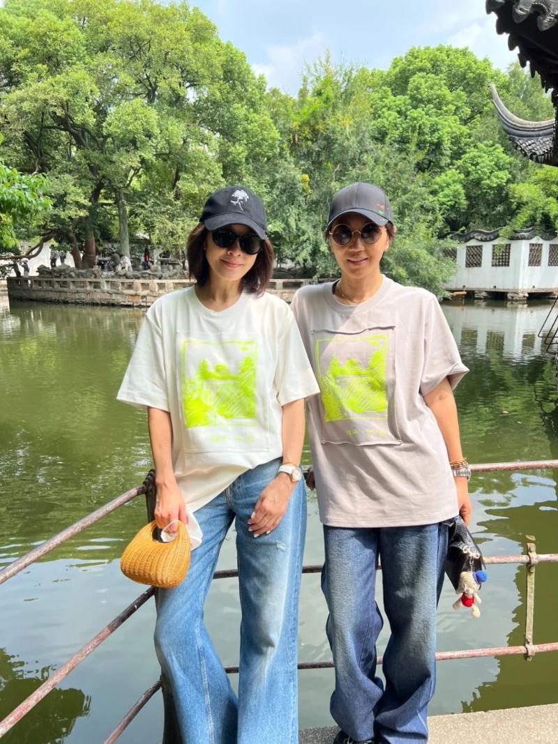 劉嘉玲頭戴黑色cap帽，身穿一件印有螢光圖案的白色短袖T恤、淺藍色牛仔長褲，身上拎住一個編織包包，非常休閑地在園林影靚相。